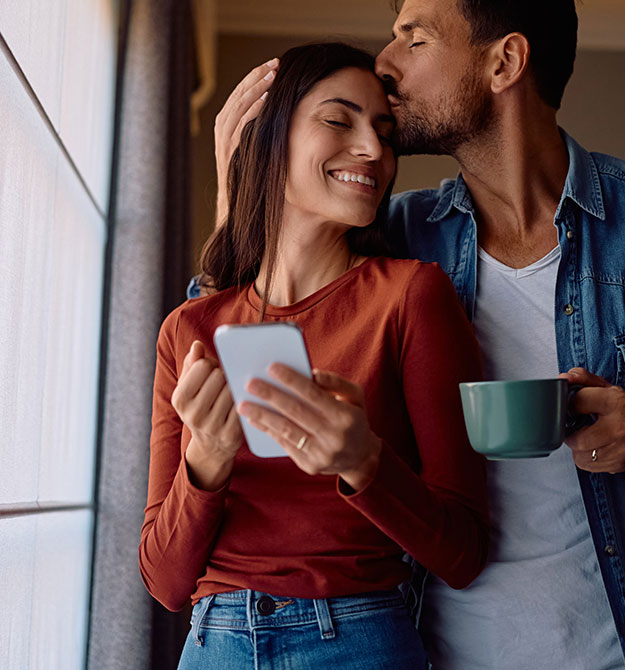 Frau hält ein Smartphone in der Hand, während ihr Mann sie auf die Stirn küsst