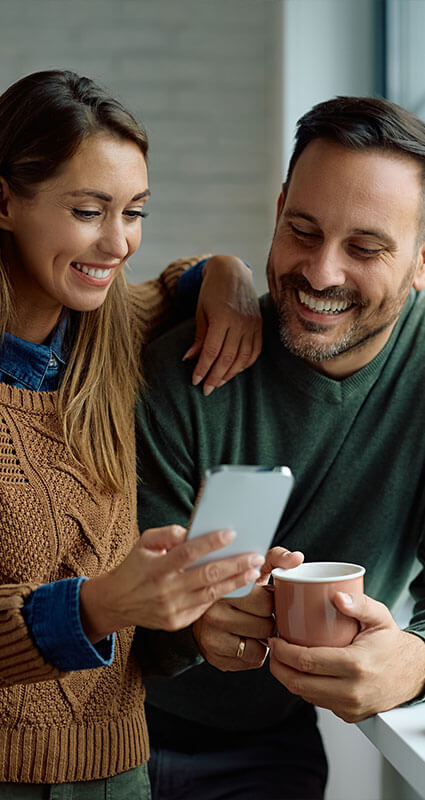 Ein Paar mit Handy und Kaffeetasse strahlt: Der 100.000-Euro-Kredit ist in trockenen Tüchern.