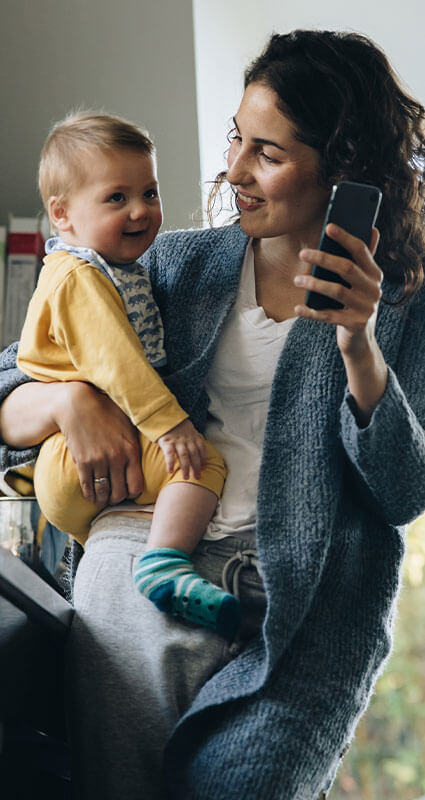 Frau informiert sich über Vorausetzungen für einen Kredit am Smartphone während sie ihr lächelndes Kleinkind auf dem Arm hält