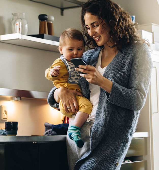 Frau hält ihr Kleinkind auf dem Arm während sie auf ihrem Smartphone Kreditangebote anschaut