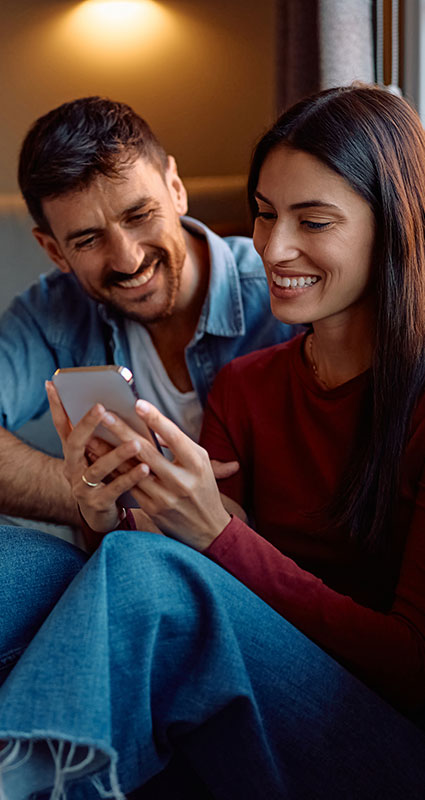 Frau informiert sich über Vorausetzungen für einen Kredit am Smartphone während sie ihr lächelndes Kleinkind auf dem Arm hält
