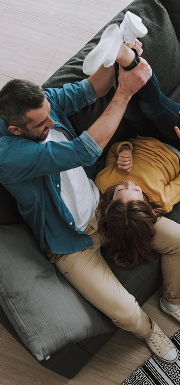 Vater sitzt auf dem Sofa und hält dabei spielerisch die Beine seiner lachenden Tochter nach oben