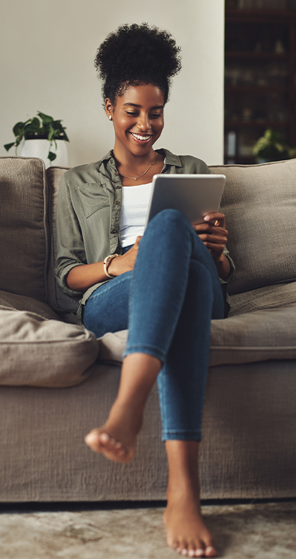 Junge Frau mit Tablet auf dem Sofa schmunzelt wegen der günstigen Prolongation ihrer Hausfinanzierung.