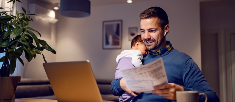 Ein Vater mit schlafendem Baby prüft am Laptop und mit Unterlagen eine Konditionsanfrage.
