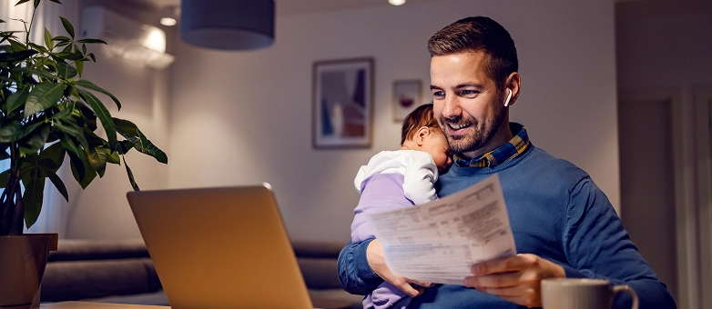 Ein Vater mit schlafendem Baby prüft am Laptop und mit Unterlagen eine Konditionsanfrage.