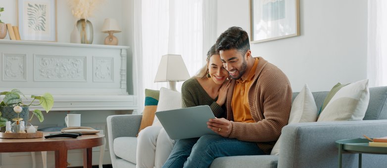 So finden Sie den passenden Ratenkredit. Ein Paar sitzt auf dem Sofa und informiert sich am Laptop über einen Ratenkredit.