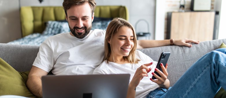 So beantragen Sie eine Ratenpause rechtzeitig. Ein Paar sitzt auf dem Sofa mit Laptop und Smartphone und informiert sich über eine Ratenpause.