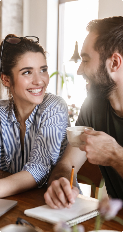 Kaffeetrinkender Mann schaut seine Frau lächelnd an, während beide am Tisch sitzen und Notizen machen